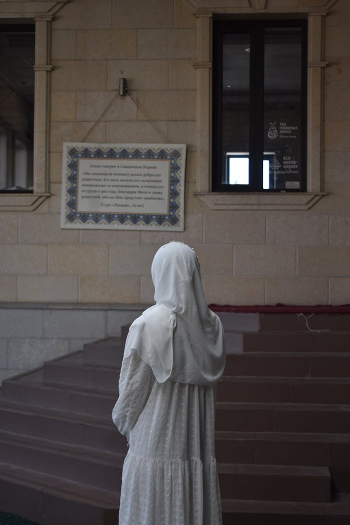 A woman in a white hijab and dress stands near a stone wall inside a building. Peaceful and contemplative scene.